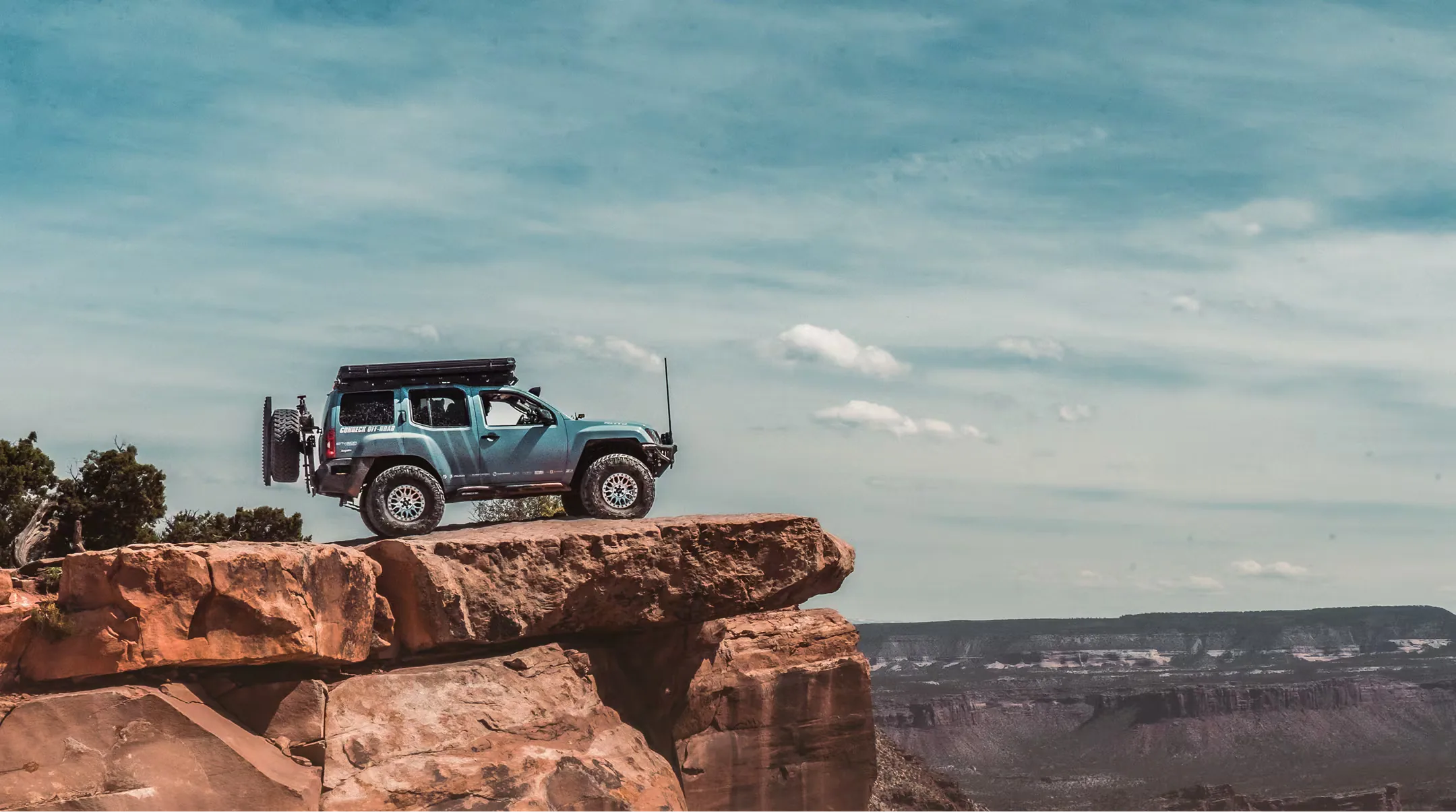 Blue off-road vehicle parked on a rocky cliff edge overlooking a canyon landscape under a blue sky.